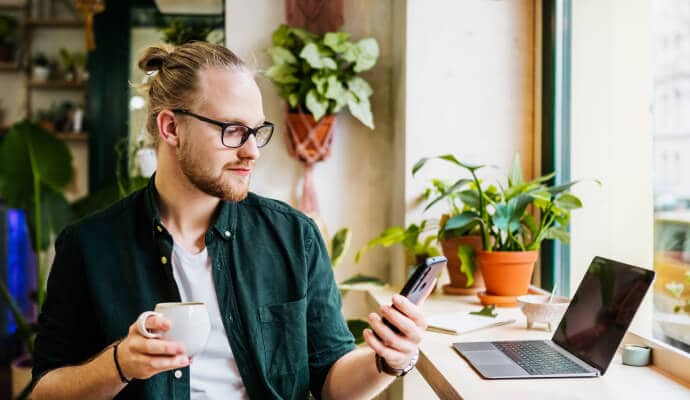 Mand, der drikker kaffe og har en mobiltelefon, der er beskyttet med Norton 360 for Mobile.